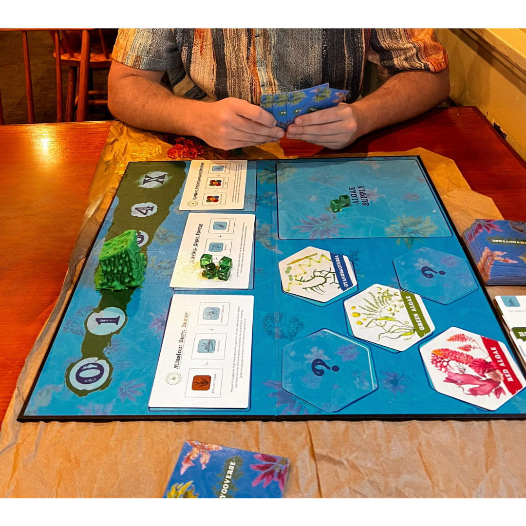 A person sitting at a wooden table playing a board game called "Phycoverse". The game board has a blue background with various cards, hexagonal tiles, and green plastic pieces representing algae. The player's hands are visible holding blue cards.
